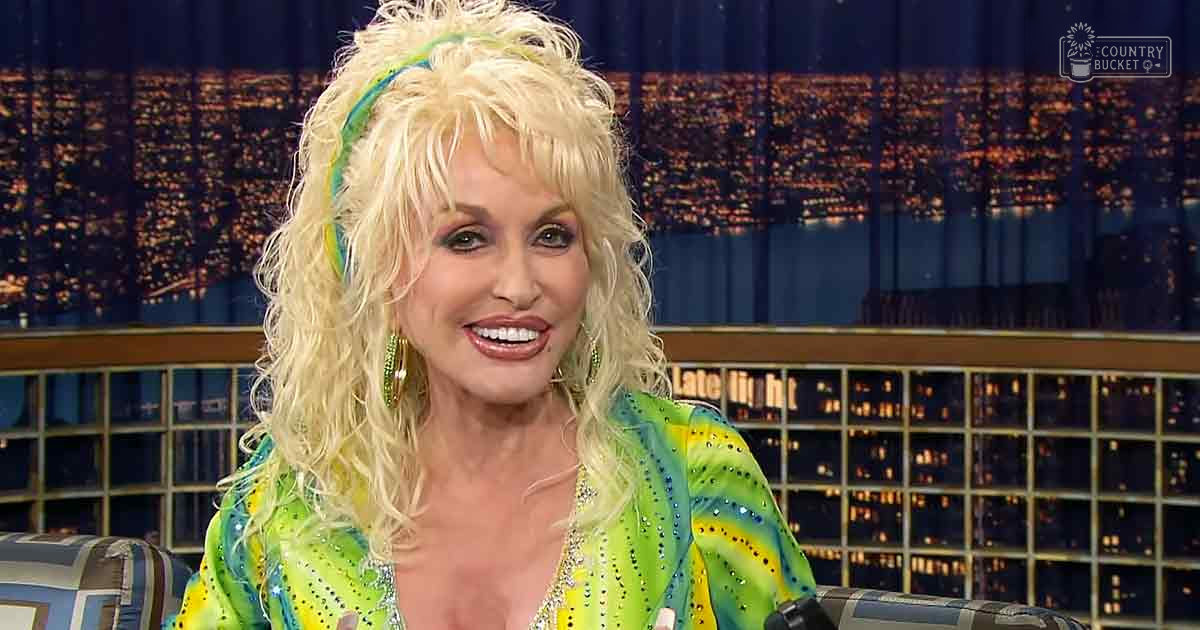 A middle-aged woman in a vibrant green and yellow patterned sequined dress on a late-night talk show set with urban night backdrop.