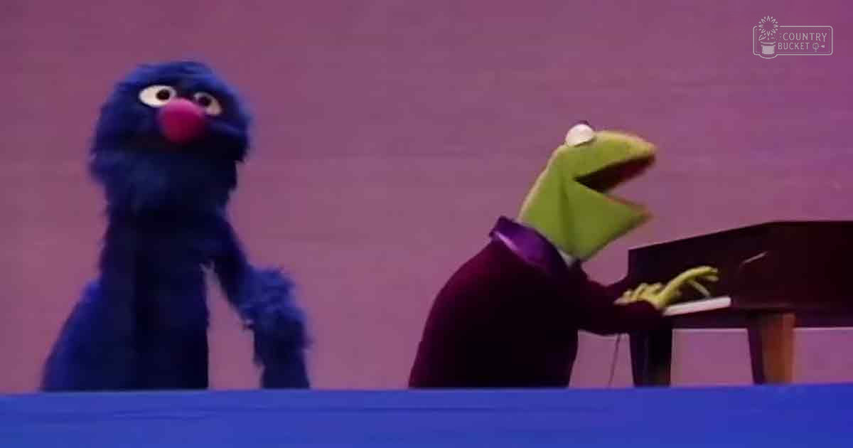 Two puppet characters, Grover in a dark blue suit with a red nose and Kermit the Frog in a dark purple jacket over a white shirt, perform on stage in an indoor studio setting.