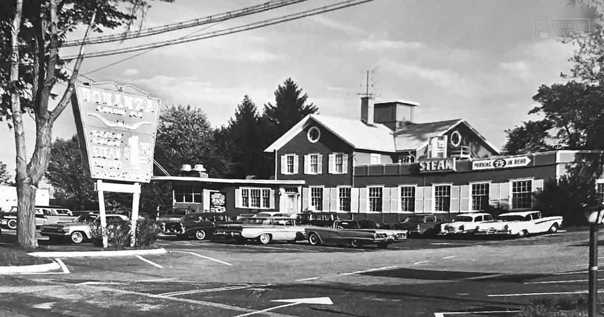 Vintage cars from mid-20th century parked outside Bollin's Steak House in a parking lot with trees and power lines, partly cloudy sky indicating daytime, season likely spring or summer.