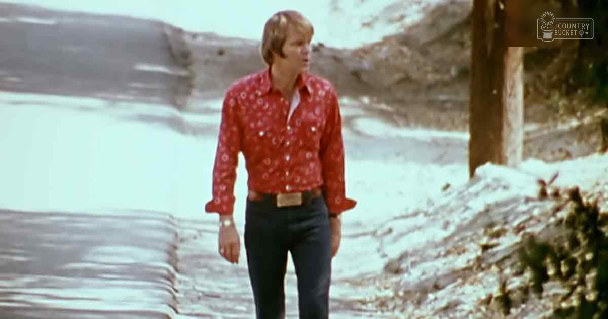 A young adult male walks along a desert path, wearing a red long-sleeved shirt with white floral patterns and beige pants. The landscape includes sparse vegetation and rocky terrain under clear midday light.