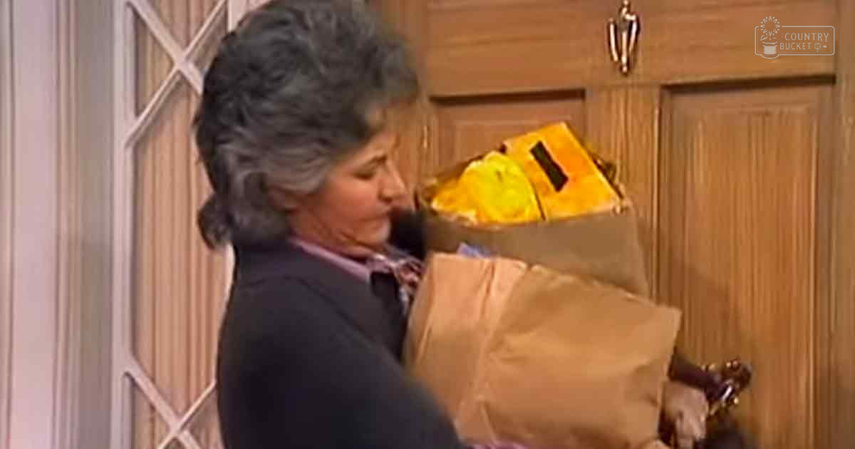 An older adult woman wearing a dark-colored long-sleeve top, holding a brown paper bag filled with groceries in an indoor domestic setting.