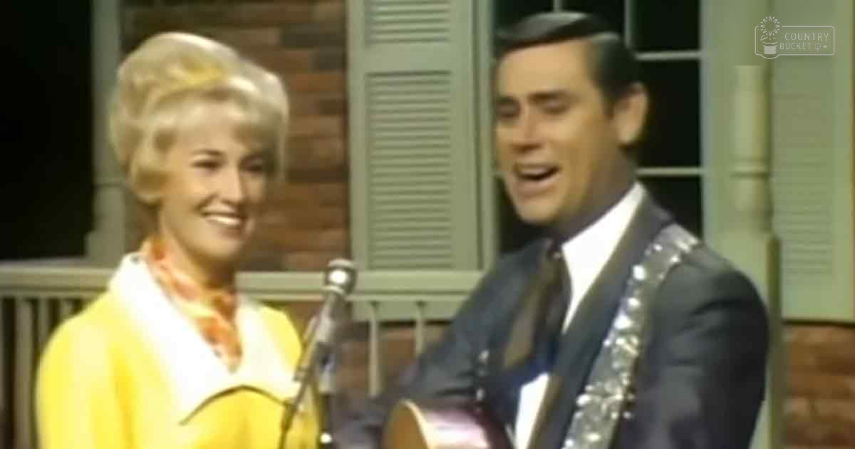 An indoor musical performance with a female in a yellow cardigan and patterned scarf, and a male in a dark suit jacket over a white shirt and tie, set against a backdrop resembling a brick wall with shutters or panels.