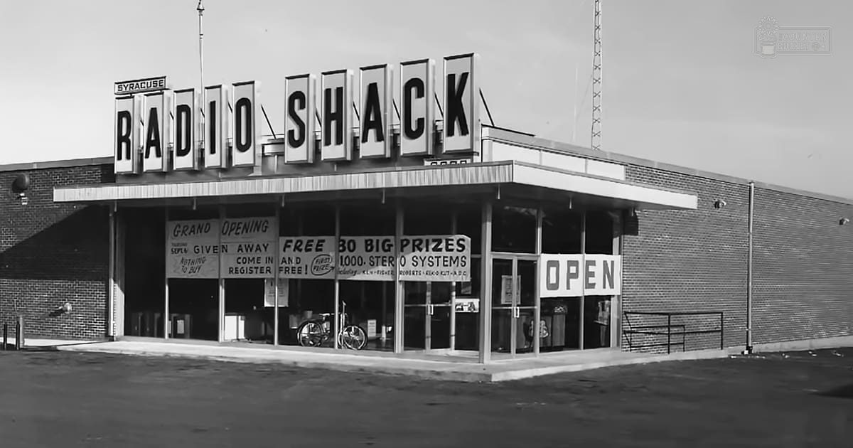 Radio Shack storefront with large signage displaying 'RADIO SHACK' and various promotional signs advertising prizes, giveaways, and free items; outdoor commercial area during daytime.