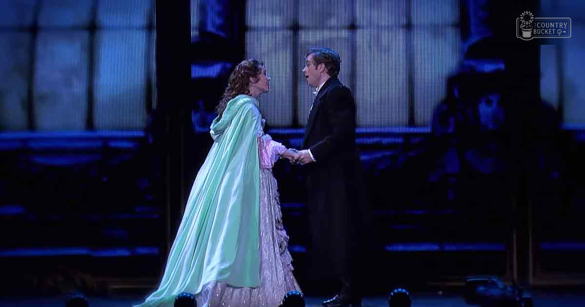 Theatrical performance scene with an adult female in a Victorian-style green cape and lace skirt, interacting with an adult male in formal attire on stage under dramatic lighting.