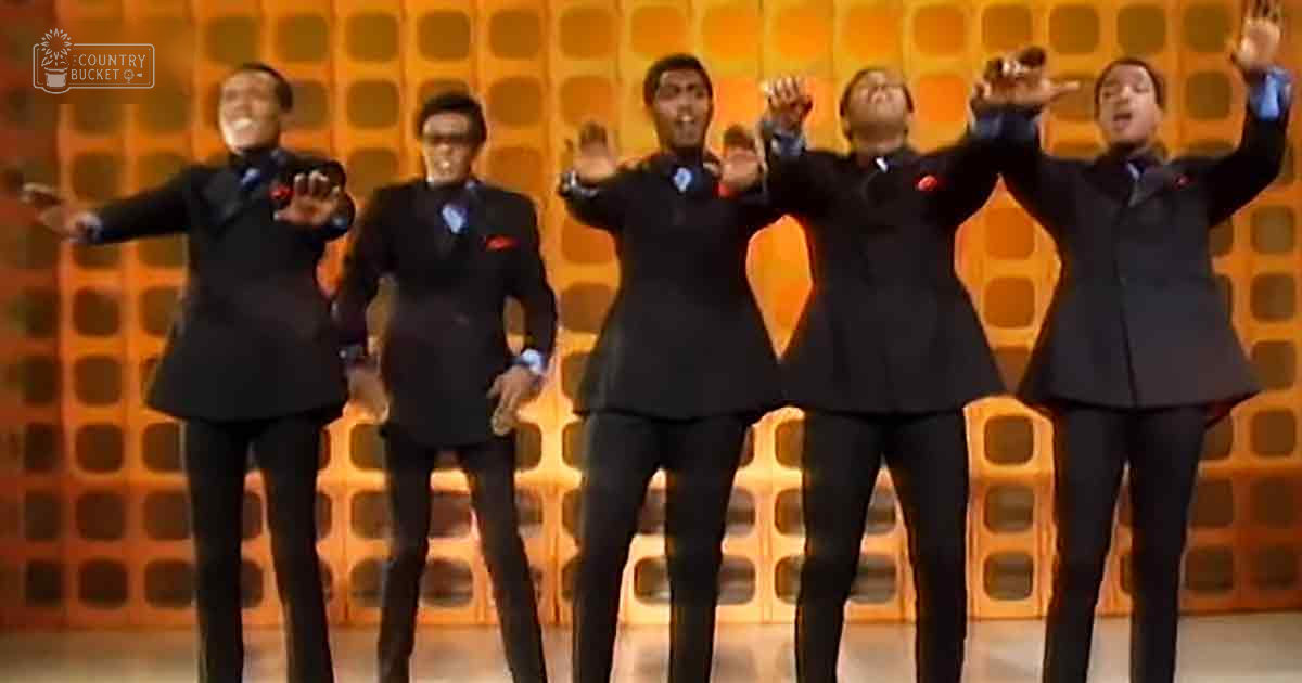 A group of adult male performers in a studio setting, dressed uniformly in classic-cut black suits with white shirts and either red or blue ties.