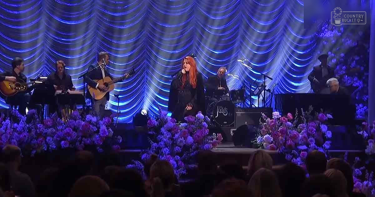 A musical performance taking place indoors with a lead singer and band playing guitars on a stage decorated with floral arrangements.