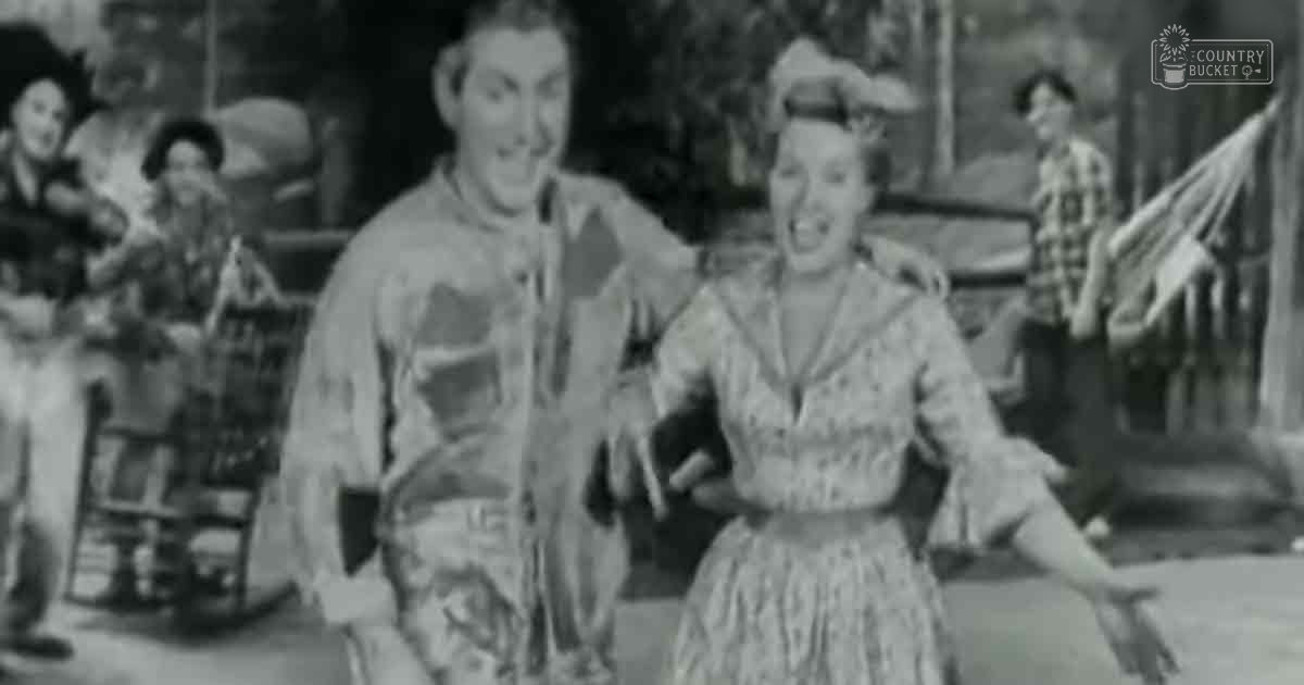 A couple dancing in a rustic rural setting with wooden structures and natural daylight; individuals wearing mid-20th-century attire suggesting spring or summer.