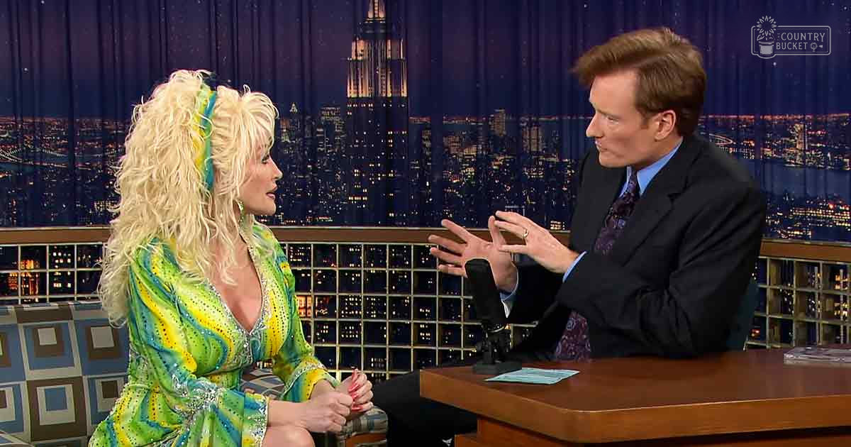 A nighttime talk show set with a woman in a green dress against an illuminated cityscape backdrop with the Empire State Building.