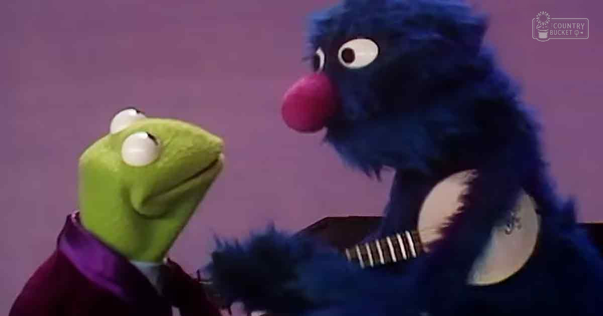 Two puppet characters, Kermit the Frog in a dark purple suit jacket with white shirt and Grover without visible clothing, set against a plain indoor background.