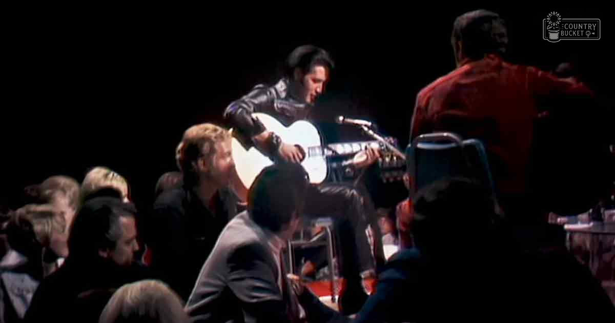A performer playing an acoustic guitar in a dimly lit indoor concert setting with seated audience members dressed formally.