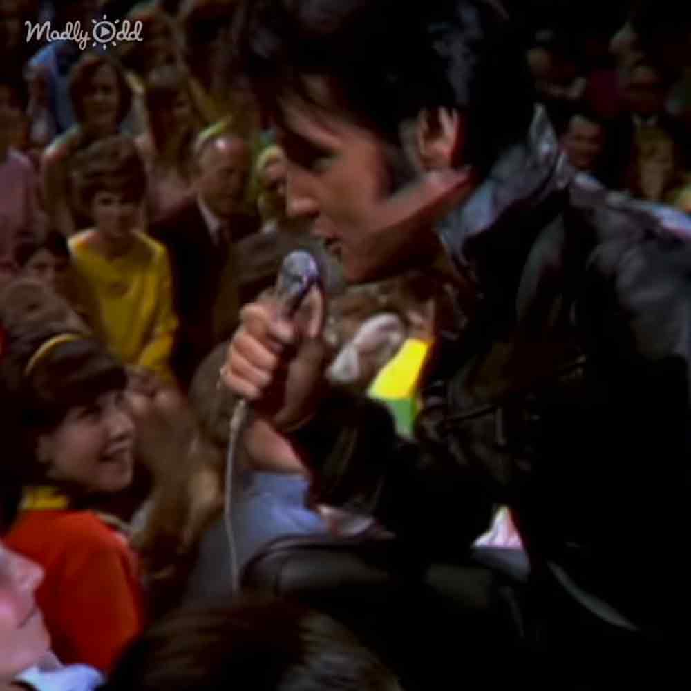 An adult male singer in a dark patterned outfit with an audience including a young female child and adults, possibly from mid-20th century attire, in what appears to be a studio or performance setting.