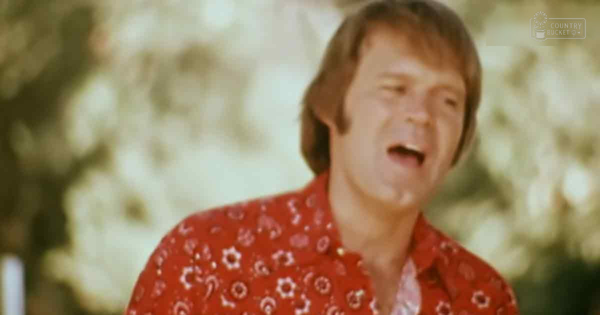 A middle-aged man wearing a red shirt with white floral designs stands outdoors amidst trees and foliage on what appears to be a bright spring or summer day.