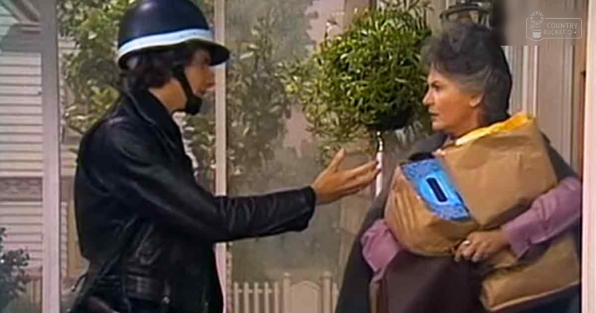 A young adult male police officer in a black leather jacket and helmet with white trim interacts with an older adult woman wearing a brown robe with blue details, indoors under natural light.