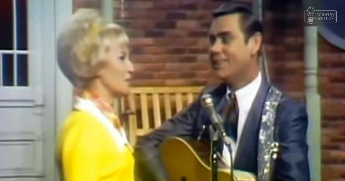 Adult male singer wearing a dark blue suit jacket and white shirt with a dark tie playing guitar in an indoor setting with a bright yellow-clad adult female, microphone nearby on a stand, brick wall background suggesting stage or performance space.