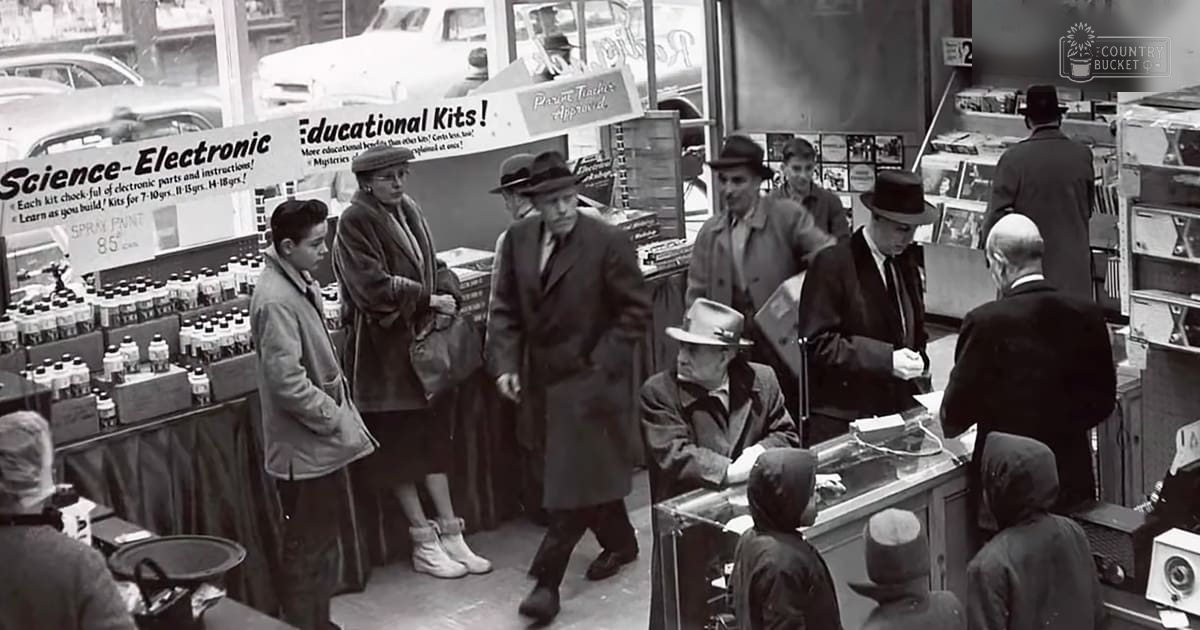 A busy mid-20th century store interior with adults and a toddler browsing products, signage reading 'Science-Electronic' and 'Educational Kits!', black and white image.