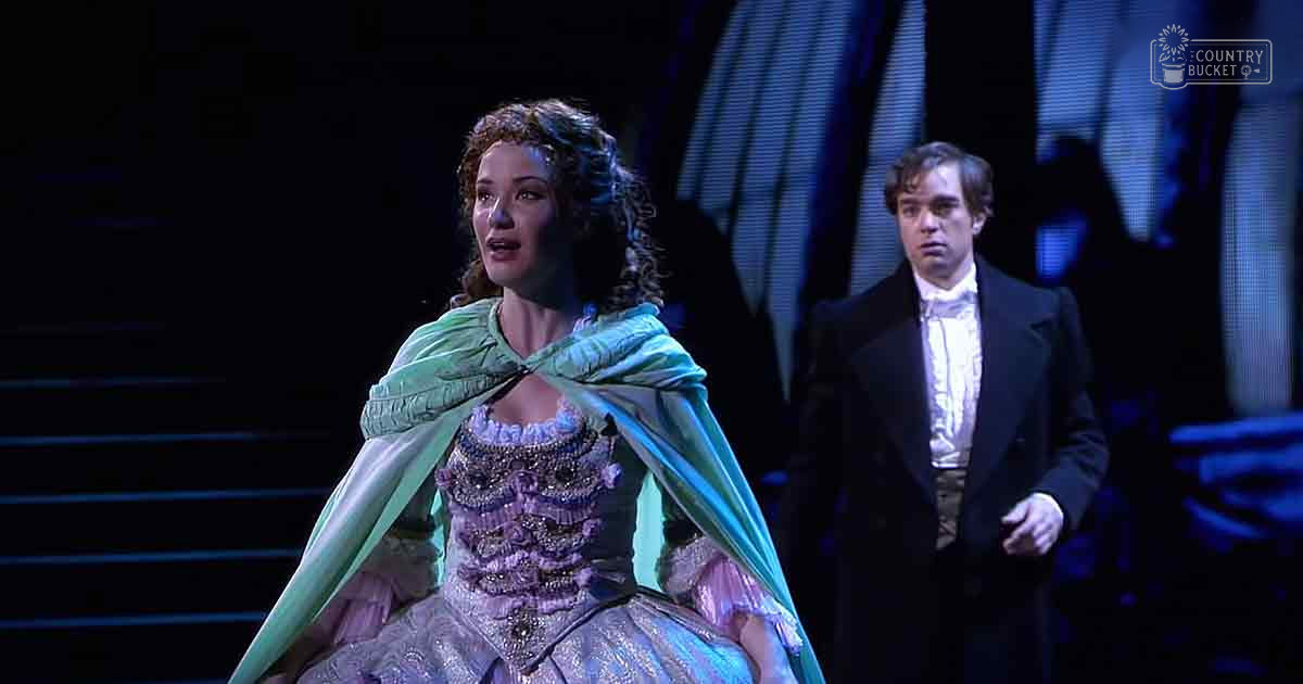 A woman wearing an ornate light purple dress with intricate beadwork and embroidery performs on stage alongside a man in a formal black suit, set against a backdrop of theatrical lighting.
