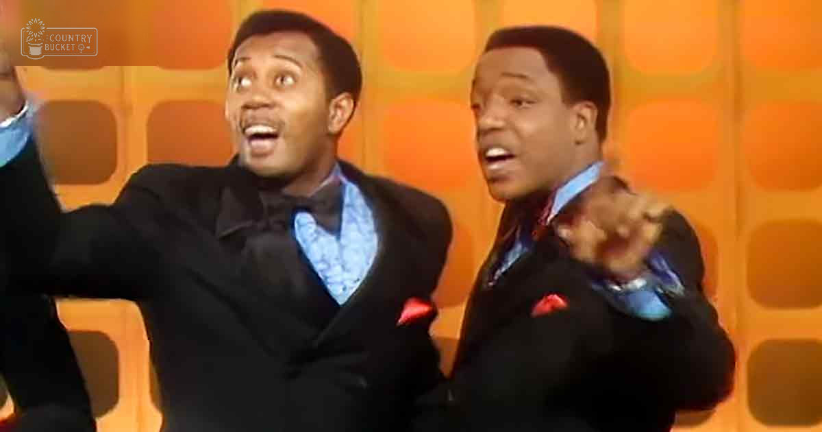 A performance scene with adult males wearing black suits, blue patterned shirts, and red pocket squares against an orange geometric backdrop.