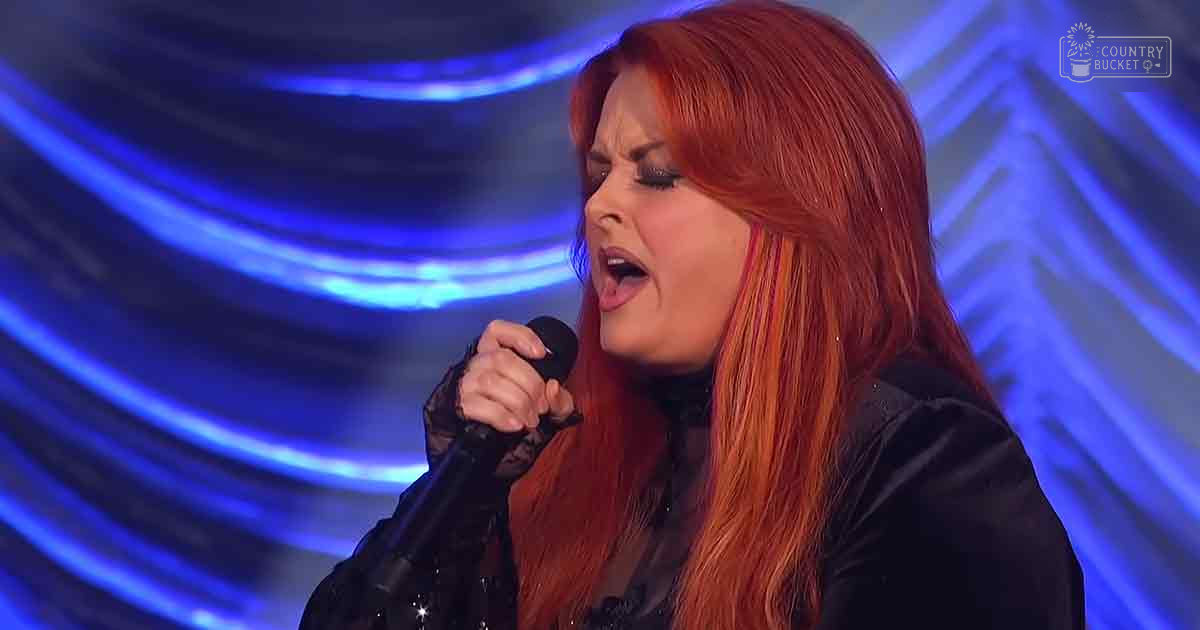 A female adult singer wearing a black lace-trimmed formal outfit, performing into a microphone against blue stage lighting with 'Madly Odd' printed in white text.