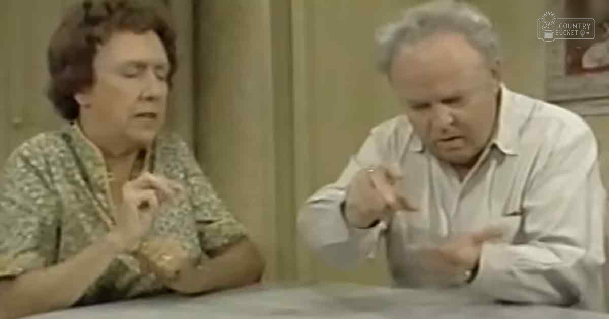 An indoor scene with a middle-aged man wearing a white button-up shirt and an older adult woman in a patterned blouse with floral designs, set against curtains and part of framed artwork.