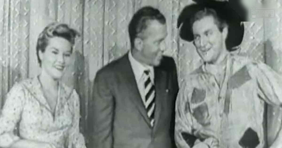 An adult male singer wearing a dark suit jacket with vertical stripes and a white shirt under it, standing indoors near two other individuals in formal attire from the mid-20th century; textured wall backdrop suggests a home or small venue setting.