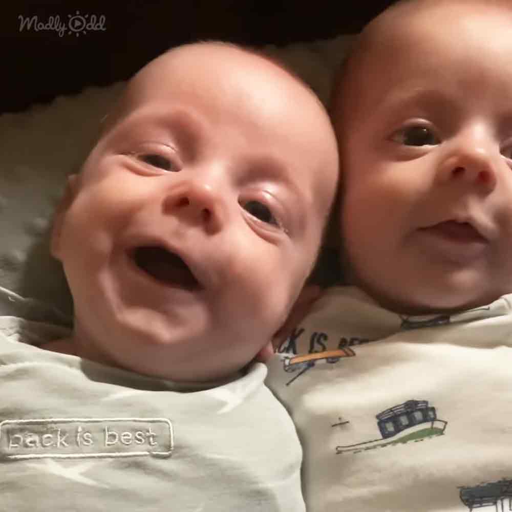 Two infants wearing light-colored onesies with a house pattern, possibly indoors at night.