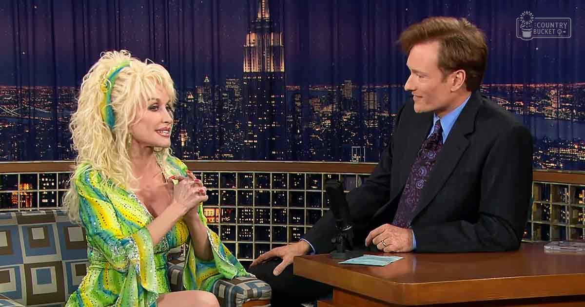 A middle-aged female in a vibrant green and yellow patterned dress with sequins, styled with large curls and colorful feathers, speaks to an adult male singer in a dark suit on a talk show set mimicking New York City at night.