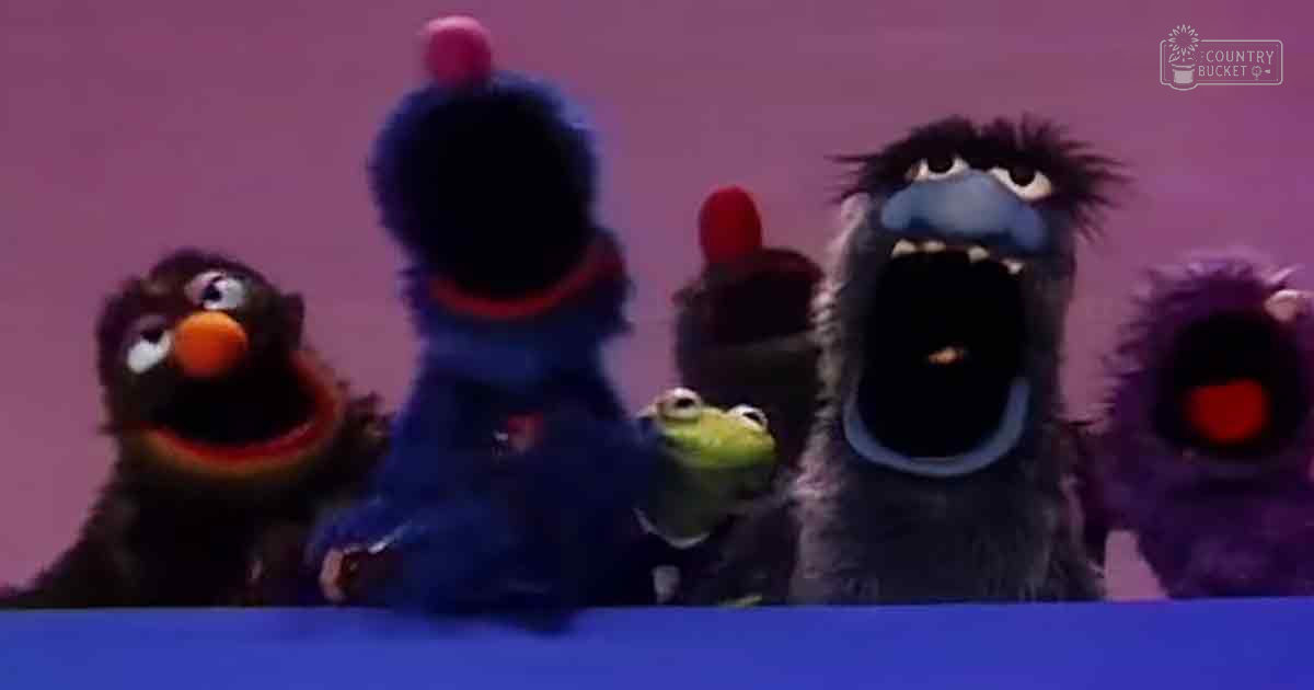 Blue puppet with a red collar and small green frog on its hand in a studio setting with other puppets, pink background, daytime lighting.