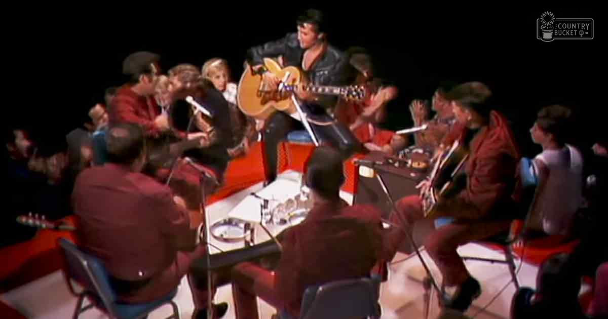 An adult male singer performing at a concert hall, surrounded by band members wearing red outfits, with stage lighting and dark curtains typical of such venues.