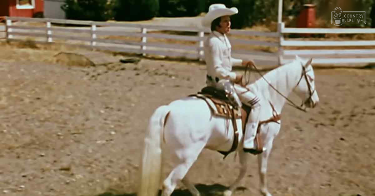An adult male wearing a white cowboy hat, long-sleeved shirt with light-colored pants rides a white horse in an outdoor equestrian arena during daytime.