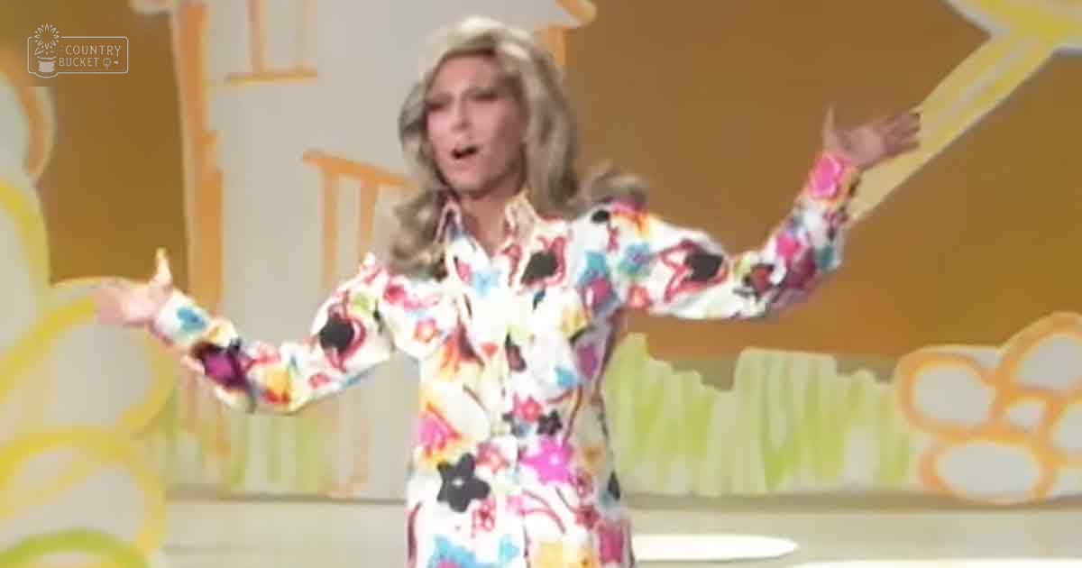 An adult female singer in a loose-fitting, long-sleeved floral dress with pink, blue, and black flowers on white from the 1960s or 70s, performing indoors under stylized yellow-green floral backdrop.