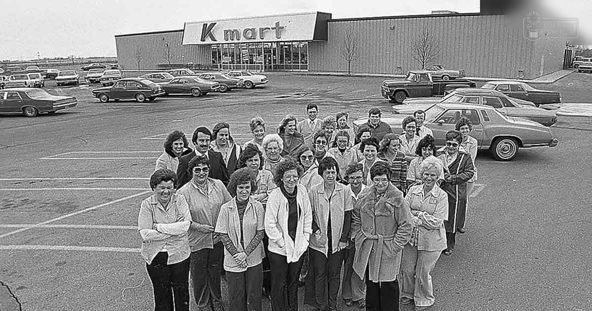 A group photo taken outdoors at a Kmart parking lot with multiple adult females wearing jackets and casual attire in late fall or early spring, under overcast daylight conditions.
