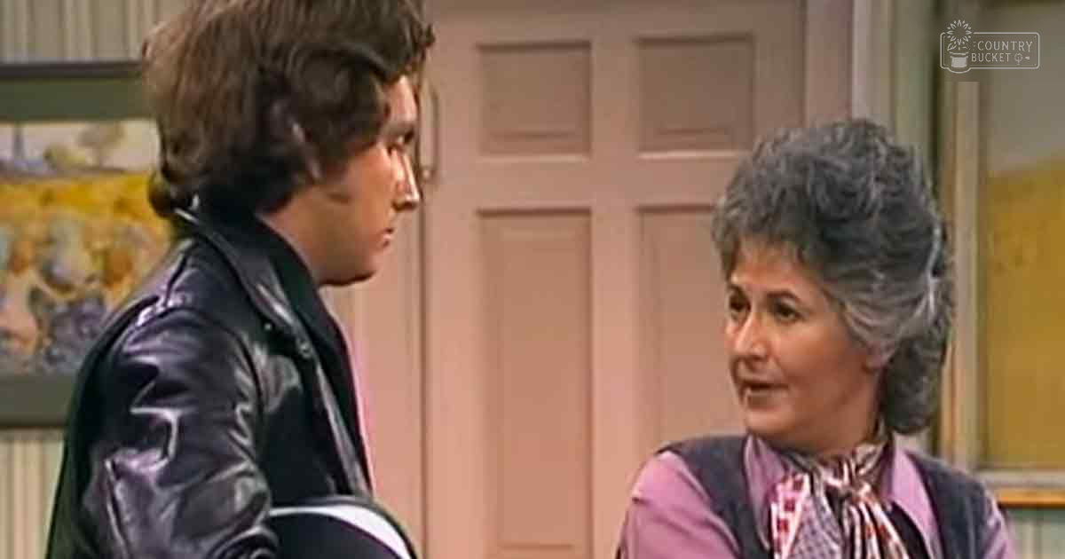 Young adult male wearing a black leather jacket with white stripes, standing indoors alongside an older adult female in a purple shirt and checkered scarf.