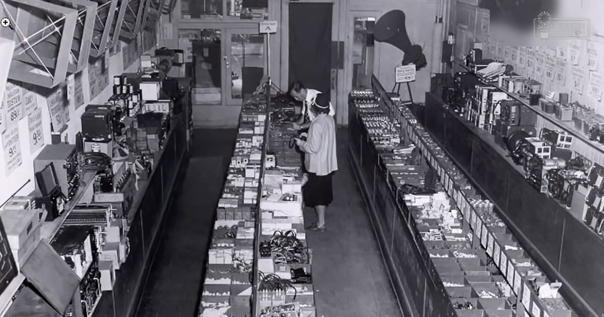 Mid-20th century store interior with shelves displaying various electronic devices, clothing, and signs indicating prices.