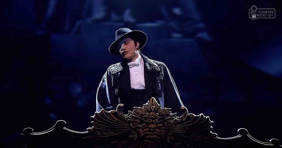 An adult male performer in a theatrical setting, wearing a black hat with a wide brim, white shirt with a bow tie, dark jacket adorned with decorative embellishments from the late 19th to early 20th century; ornate stage prop visible at the bottom of the frame.