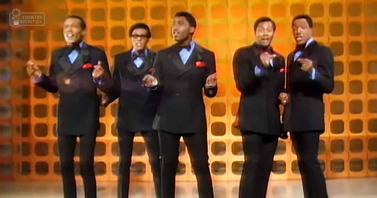 Group of adult male performers in a studio setting wearing dark suits with light blue shirts and red pocket squares, mid-20th century style.