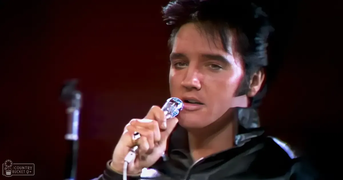 Adult male singer wearing a dark, fitted shirt holding a metallic microphone on an indoor stage with blurred background elements.