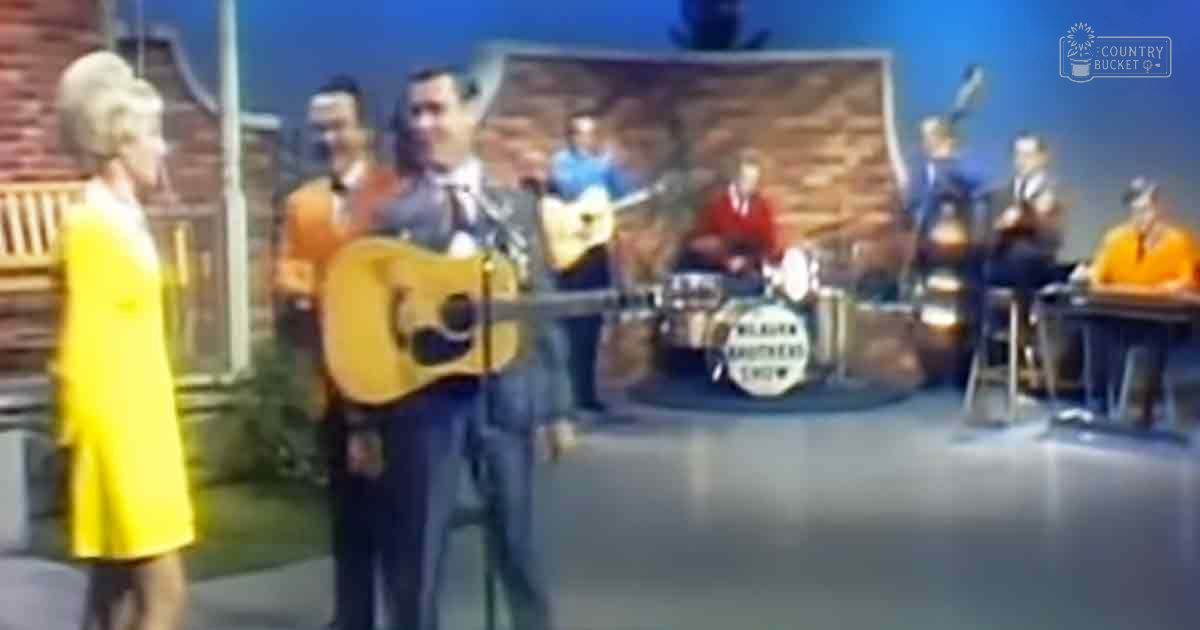 An adult male musician plays guitar at an outdoor music show, with a female audience member in a yellow dress and band members in colorful shirts against a brick wall backdrop.