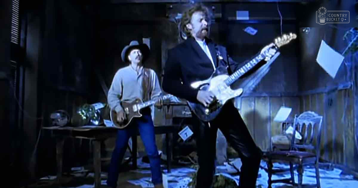 An indoor musical performance with two adult male performers wearing casual and formal attire respectively; dim blue-toned lighting creates a moody ambiance in what seems like a basement setting.