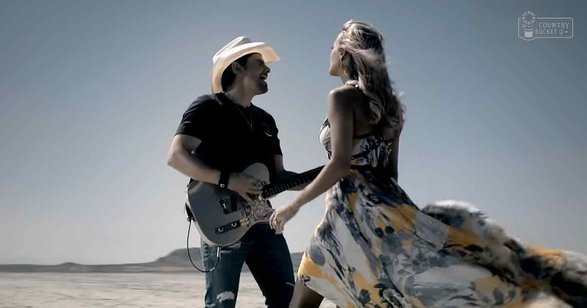 A couple in a desert setting; male adult wearing a black t-shirt with logo, blue jeans with rips, and cowboy hat; female adult in a sleeveless floral dress, holding a guitar under bright daylight.