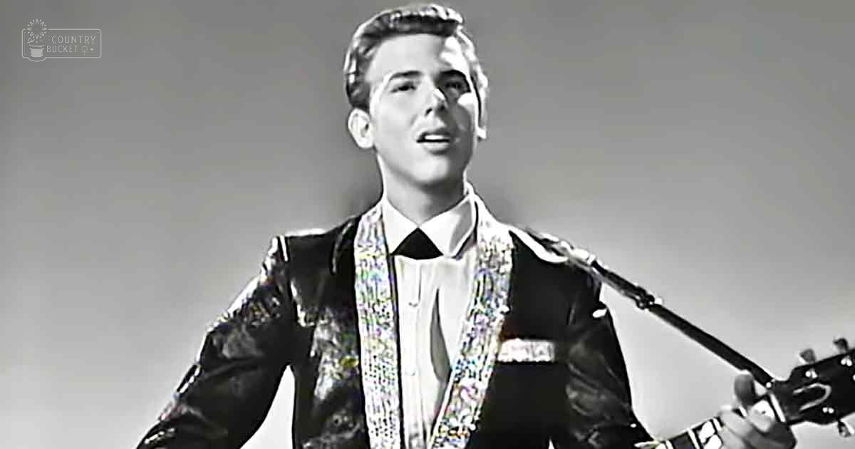 A young adult male in a relaxed fit leather jacket with decorative embellishments, white collared shirt underneath, black and white attire, indoors with guitar visible.