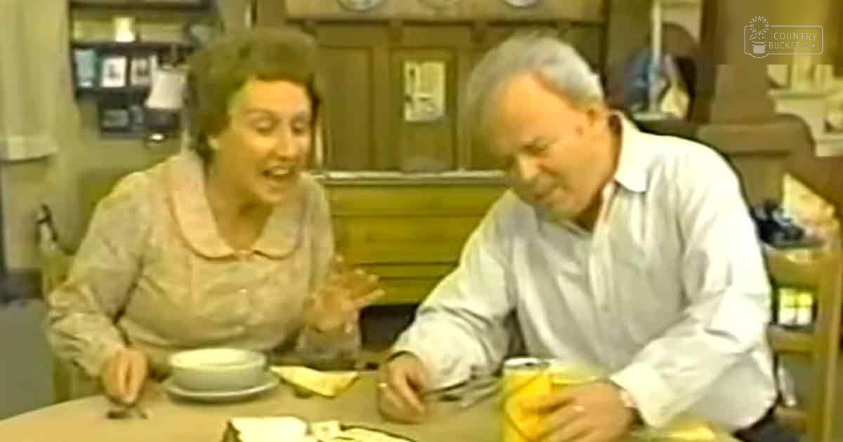 Middle-aged to older adult female wearing a light-colored, possibly floral or paisley patterned blouse with long sleeves at an indoor diner setting with vintage decor. Older adult male in similar attire sits beside her.