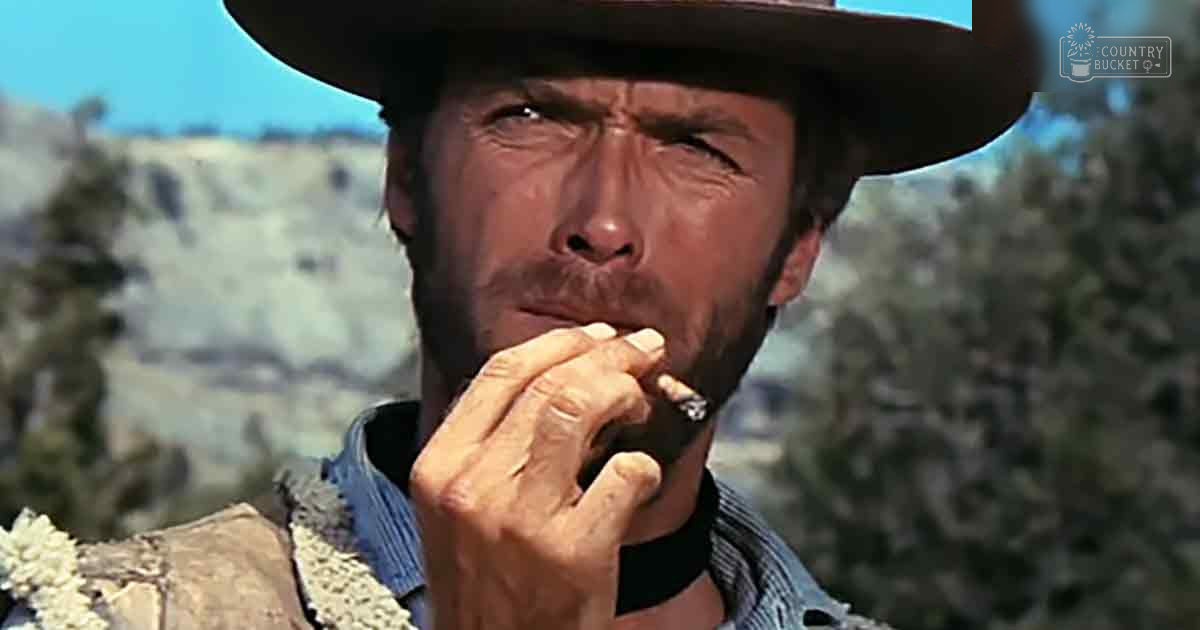 A middle-aged man wearing a wide-brimmed cowboy hat and plaid shirt in a Western landscape with sparse vegetation and mountains.