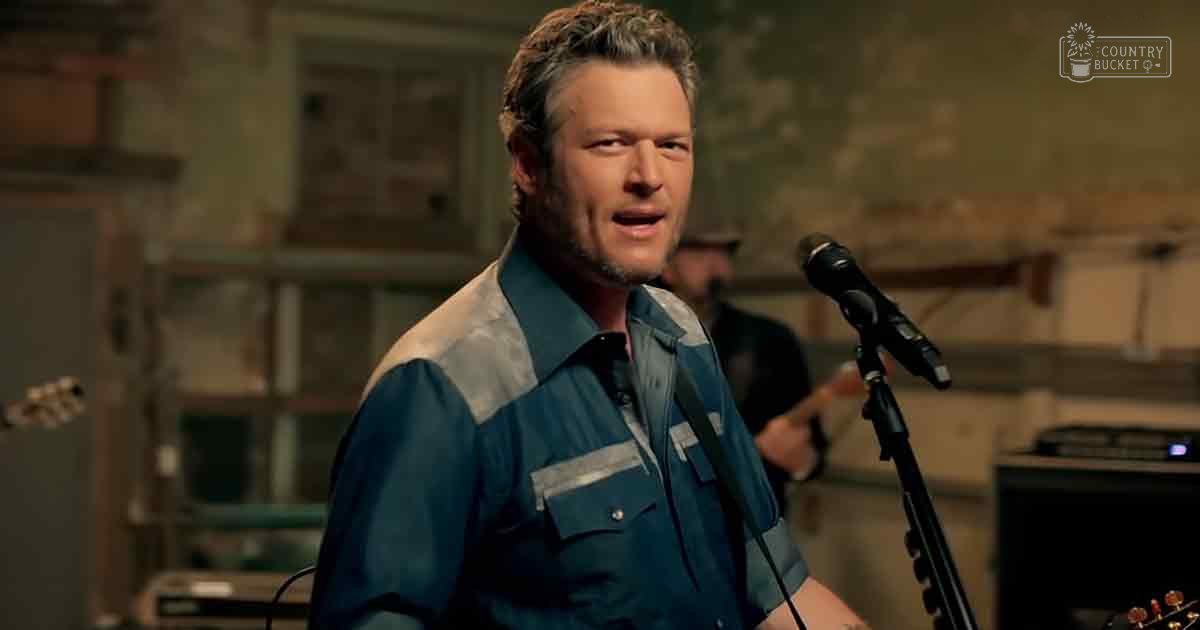 A middle-aged to older adult male in a music studio or performance space with a blue and gray patterned shirt, surrounded by musical instruments and a rustic setting.