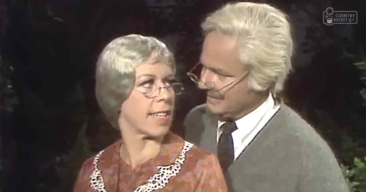 An older adult couple dressed in vintage attire in a dimly lit setting with muted earthy tones.