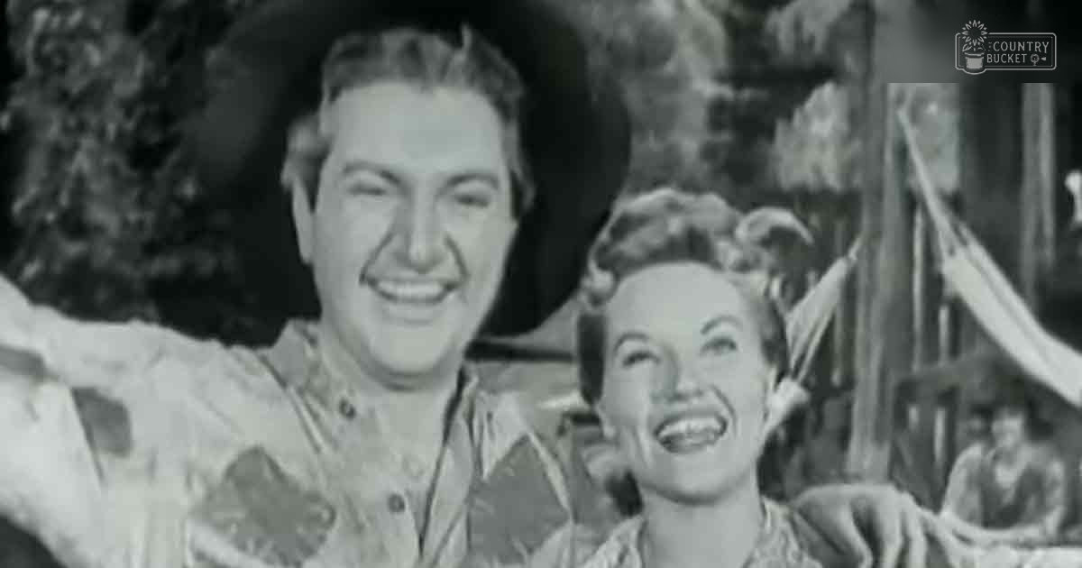 A man and a woman in tropical outdoor setting with 1940s-style clothing under bright daylight; man wears leaf-patterned shirt, woman has fitted dress with puffed sleeves.