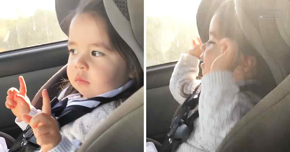 A toddler in a white sweater with a textured pattern, black bow tie or ribbon around her neck, and fingerless gloves sits in a car seat inside a vehicle. Sunlight streams through the window, suggesting daytime in spring or summer.