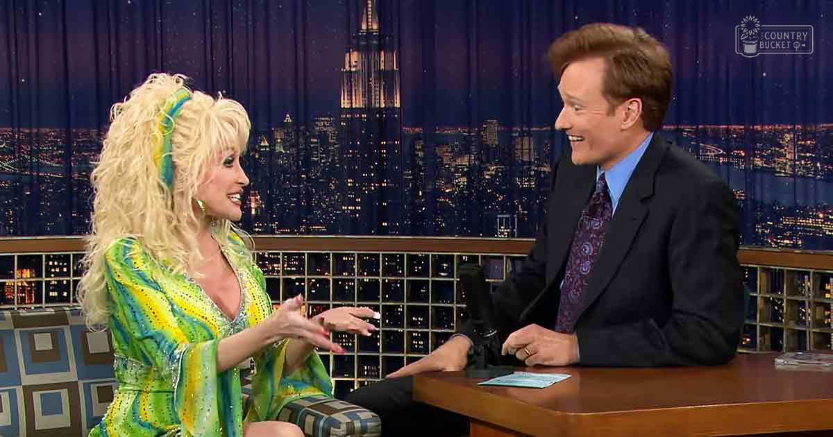 Two adults, a female in a green patterned dress and male in a dark suit, seated for an interview against a cityscape backdrop at night.