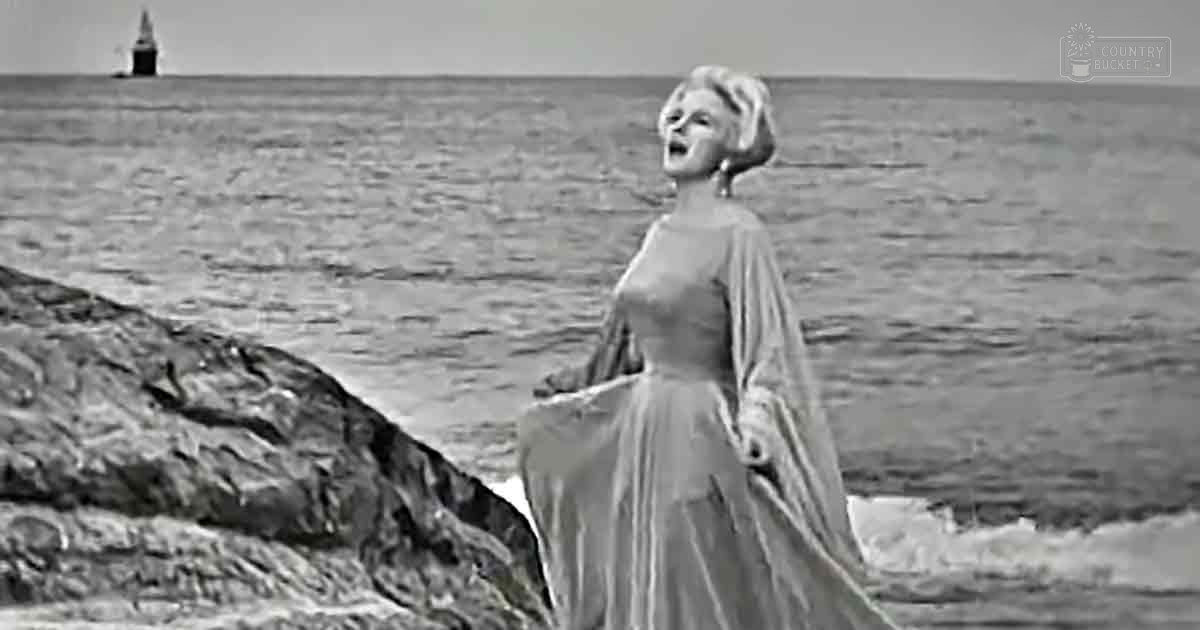 A woman singing in front of an ocean setting with a lighthouse visible in the distance.