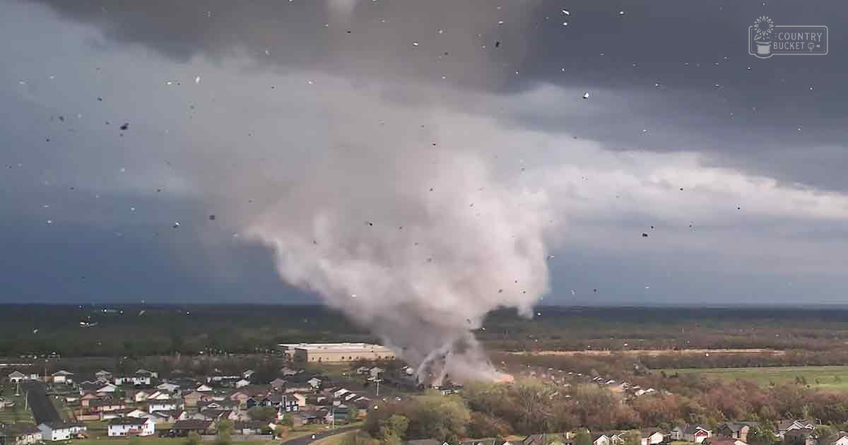 A suburban neighborhood scene with a large plume of smoke rising over houses, trees, and fields under an overcast sky during daytime.