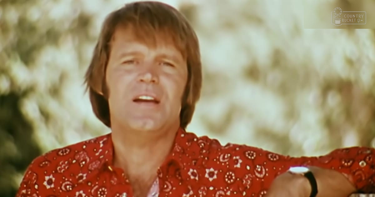 A middle-aged man wearing a red, white, and black patterned long-sleeved shirt with a collared neckline stands outdoors amidst green foliage.
